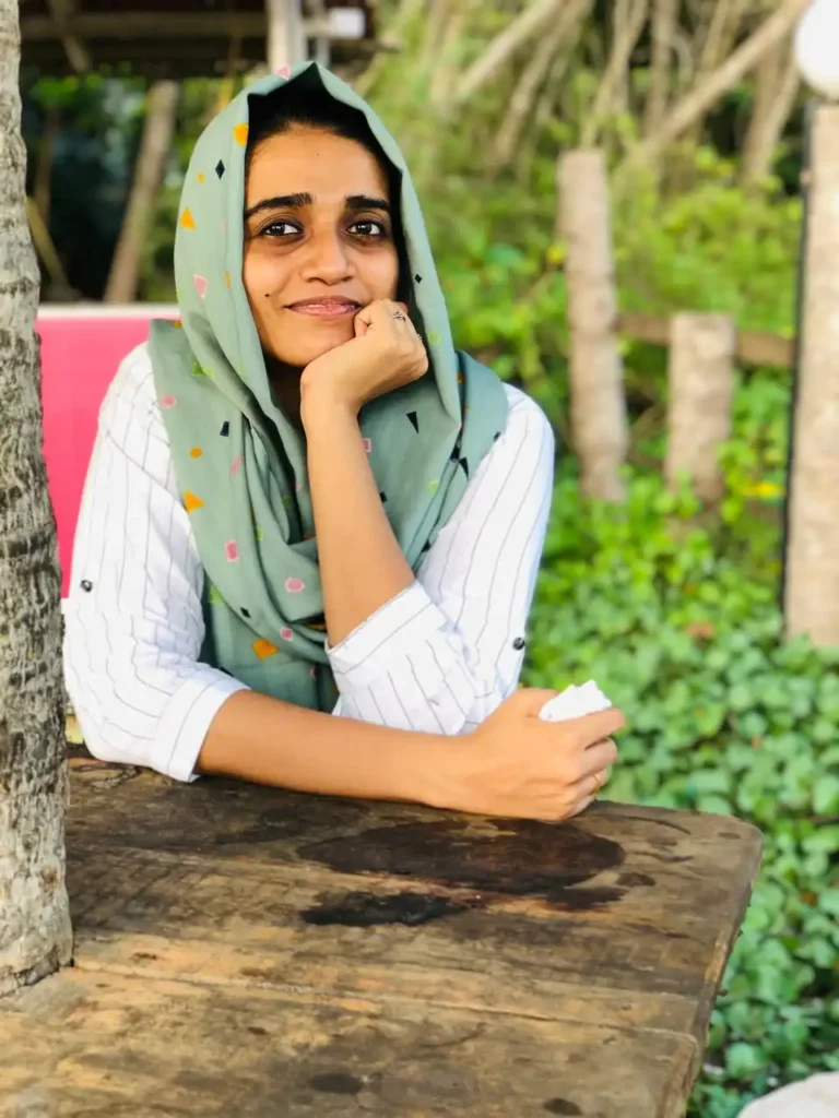 A smiling woman wearing a light green hijab and white shirt, resting her chin on her hand at a wooden table outdoors, with lush greenery and trees in the background. This image could represent a digital marketing expert in Alappuzha.